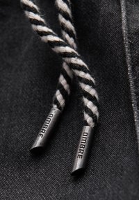 Tissu en denim noir avec des cordons de serrage torsadés noirs et blancs, ornés d'embouts métalliques gravés "OMARE". Gros plan mettant en valeur la texture.