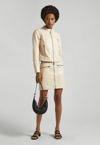 Chaqueta de cuero crema con cremalleras, combinada con una falda de cuero a juego. Presenta un bolso negro y tacones negros con tiras. Textura suave.