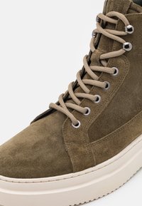 Baskets montantes en suède kaki avec des lacets beiges, œillets en métal et semelle en caoutchouc blanche, présentant une texture lisse et des détails de couture.