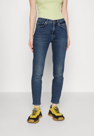 Persoon in slim-fit blauwe jeans, een limoen groene mouwloze top en stevige gele en zwarte sneakers, staand tegen een witte achtergrond.