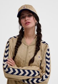 Beige Zip-Jacke mit einem dezenten Muster, marineblauen und cremefarbenen Zickzackstreifen an den Ärmeln. Passende beige Mütze mit einem gestickten Logo.