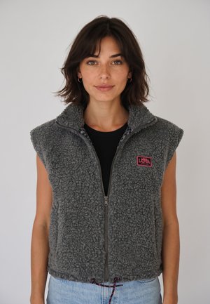 Femme aux cheveux courts et foncés portant une veste grise sans manches en tissu duveteux zippée et un jean bleu clair sur un fond blanc uni.