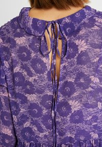 Lödereinen kukkamekko, jossa on aaltoileva kaulus ja takana solmittava kiinnitys, pehmeä, teksturoitu kangas ja hienovaraiset vaaleanpunaiset kukkakuviot.