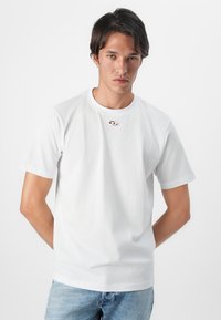 Tricou alb din bumbac, cu mâneci scurte, guler rotund și un mic logo accentuat aproape de guler. Modelul poartă blugi de culoare albastru deschis.