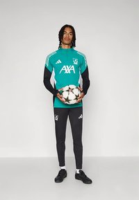 Νεαρός άνδρας αθλητής σε τιρκουάζ και μαύρη αθλητική ένδυση κρατάει μια άσπρη και μαύρη μπάλα ποδοσφαίρου, στέκεται μπροστά σε ένα απλό ανοιχτό φόντο.