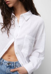 White linen button-up shirt with an open placket, single chest pocket, and relaxed fit. Paired with high-waisted blue denim jeans.