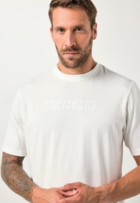 Bearded man s tetováním na ruce, oblečený v bílém tričku "PROJECT UNLMTD", stojící s dlaněmi spojenými proti jednobarevnému pozadí.