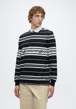 Młody mężczyzna z kręconymi włosami, ubrany w czarno-białą, paskowaną, długorękawową koszulkę rugby z napisem „tommy jeans” oraz beżowe spodnie, stojący nieruchomo.