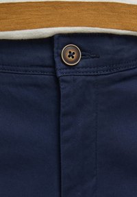 Pantalons bleu marine avec une texture lisse, dotés d'une fermeture à bouton marron et de surpiqûres. Une chemise à rayures horizontales est partiellement visible au-dessus.