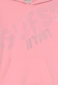 Jasnoróżowa bluza z kapturem z pojemną kieszenią z przodu, zdobiona metalicznymi literami z kryształkami, które w odważnym projekcie głoszą "GUESS // 1981".