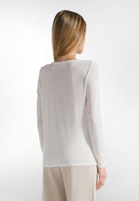 Top a maniche lunghe, semi-trasparente, di un leggero beige, con una vestibilità rilassata. Presenta un collo rotondo e una texture morbida, abbinato a pantaloni in tonalità chiare.