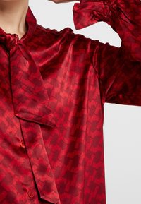 Rote Bluse aus einem seidigen Material mit einem allover abstrakten Muster, einer gebundenen Kragenpartie und einer knopfgeschlossenen Vorderseite mit gesammelten Bündchen.
