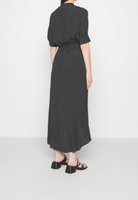 Kobieta w czarnej midi sukience w białe kropki i czarnych sandałach na obcasie, stojąca na tle białego tła, widziana z tyłu.