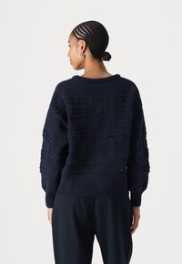 Maglione lavorato a maglia blu navy con motivi testurizzati, spalle scese e polsini a costine. La parte posteriore presenta un design a maglia aperta nella sezione superiore.