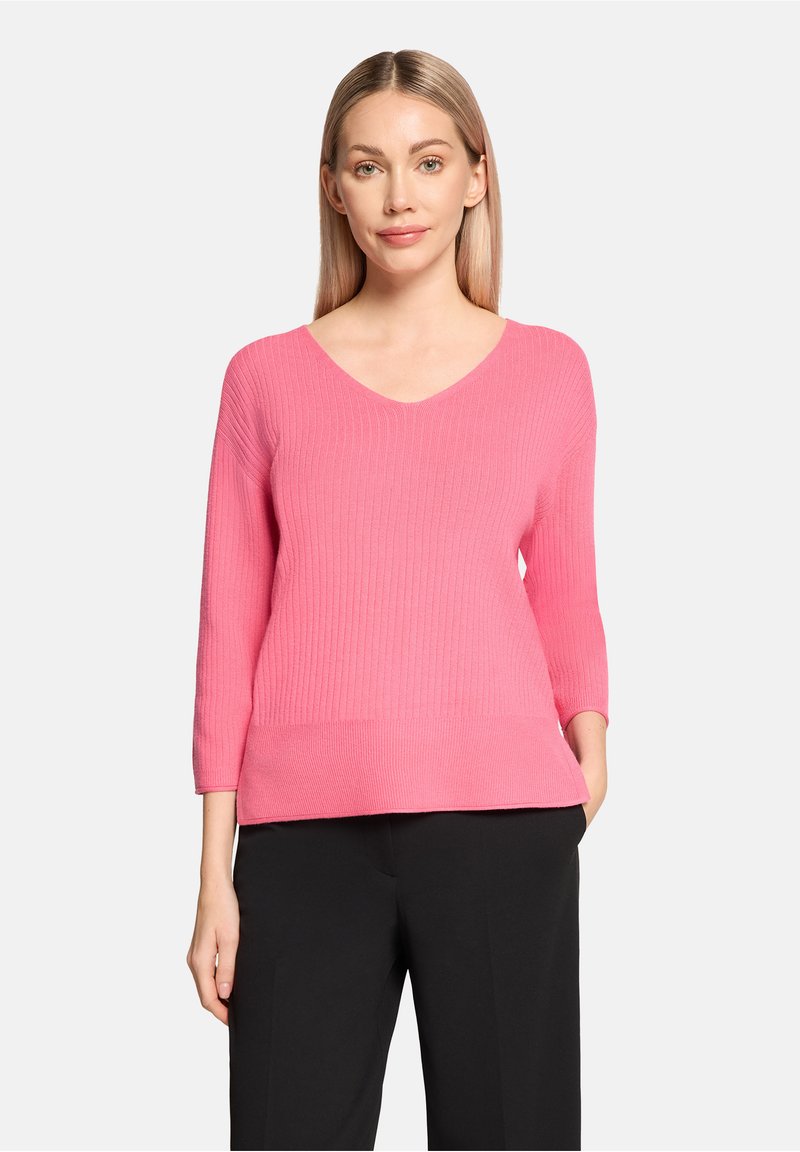 Pull en maille côtelée rose avec un décolleté en V et des manches trois-quarts, associé à un pantalon noir. Le matériau semble doux et texturé.