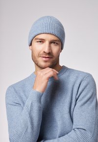 Cappello di lana azzurro chiaro a maglia con texture a coste, abbinato a un maglione a coste azzurro chiaro dello stesso colore. Uomo in posa con la mano sul mento.