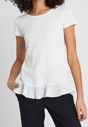 Femme portant un haut blanc à manches courtes avec un ourlet à volants et un pantalon noir à fines rayures, posant avec une main dans la poche.