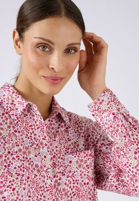 Camisa de flores en rosa claro con patrones intrincados en rojo, púrpura y verde; cuenta con cuello, botones y un bolsillo en el pecho.