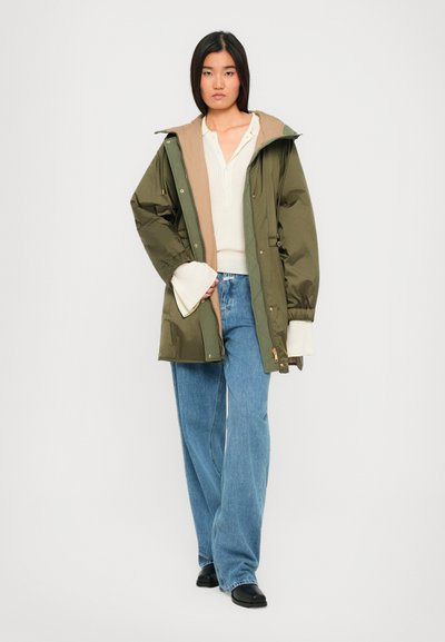 Woman standing wearing olive green hooded parka, cream knit sweater with flared sleeves, blue wide-leg jeans, and black shoes on white background.