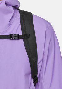 Lila utomhusjacka med svart axelrem, med en strukturerad yta och en plastspänne. Nära bild av axelområdet.