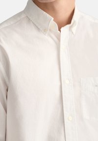 Bílá bavlněná košile na knoflíky s klasickým límečkem, přední kapsou a jemnou texturou. Knoflíky jsou bílé, což umocňuje čistý design.