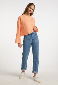 Top court à manches longues et côtelé en pêche, associé à un jean taille haute bleu clair. Le modèle porte des baskets blanches et des boucles d'oreilles multicolores.