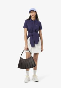 Camisa polo azul marino, falda blanca plisada, gorra azul clara, bolso negro plisado y zapatillas beige con acentos de color, sobre un fondo blanco.