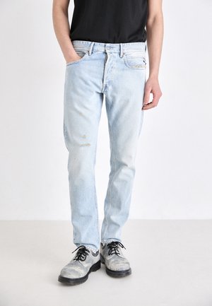 Osoba ubrana w jasnoniebieskie znoszone dżinsy, czarną koszulkę i szare sznurowane buty, stojąca z jedną ręką w kieszeni.