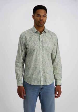 Mann trägt ein hellgrünes, langärmliges Hemd mit Blumenmuster und blaue Jeans, steht vor weißem einfarbigem Hintergrund.