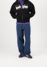 Sweat à capuche noir zippé avec texte graphique coloré, porté sur un T-shirt violet. Jean bleu ample et baskets bleu marine avec trois bandes complètent la tenue.