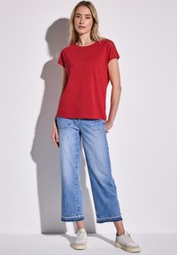 Rød kortærmet t-shirt parret med lyseblå vide jeans. Afslappet stil med en løs pasform, vist med neutrale sneakers.