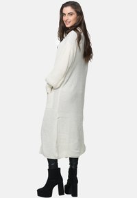 Cardigan lungo bianco a maglia con tasche laterali, caratterizzato da una vestibilità ampia e texture a costine, abbinato a stivali neri con zeppa.