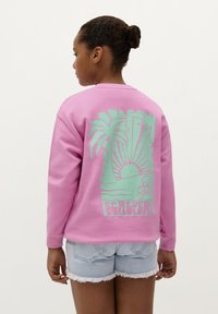 Felpa in pile rosa con maniche lunghe, caratterizzata da un design di una palma verde e di un sole sul retro. Abbinata a pantaloni shorts in denim.