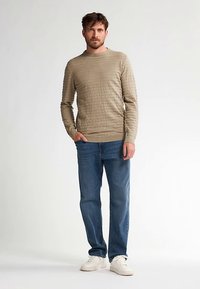 Beiger, strukturierter Pullover mit rundem Ausschnitt, kombiniert mit blauen Jeans und weißen Sneakern. Das Model steht vor einem schlichten Hintergrund.