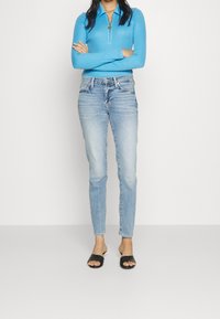 Camiseta de manga larga turquesa con cremallera, jeans ajustados de un azul claro y sandalias negras tipo slide. Conjunto casual con una textura suave y un corte ajustado.