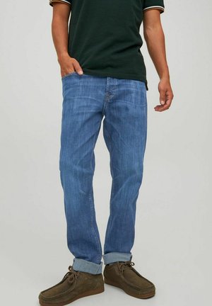 Jeans in denim blu con taglio dritto e orli arrotolati, caratterizzati da un leggero sbiadimento e dettagli di cucitura. Indossati con scarpe in suede marroni.