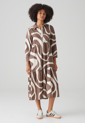 Robe chemise à motifs marron et blanc avec des manches longues, un col classique et une jupe à volants. Associée à des baskets blanches.