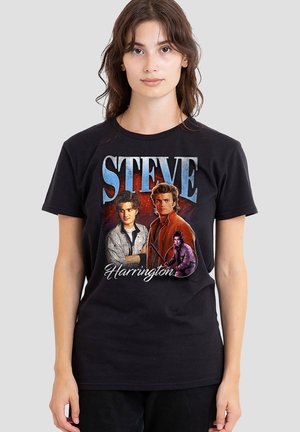 Junge Frau trägt ein schwarzes T-Shirt mit drei farbigen Porträts von Steve Harrington und dem Text "STEVE" und "Harrington".