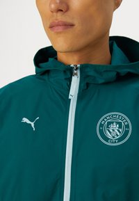 Giacca impermeabile color teal con cappuccio, dotata di una cerniera blu chiaro e di un logo Puma bianco. Presenta un emblema rotondo del club Manchester City.