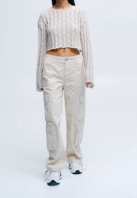 Pull court blanc en maille avec motif texturé. Pantalon cargo beige avec poches latérales. Baskets blanches et noires. Fond uni.