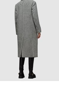 Schwarz-weiß karierter Hahnentritt-Mantel mit langer Silhouette, seitlichem Schlitz und großem Kragen. Kombiniert mit eleganten schwarzen kniehohen Stiefeln.