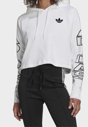 Femme portant un hoodie court blanc avec un logo noir et des inscriptions sur les manches, associé à un pantalon noir taille haute avec des bandes latérales.