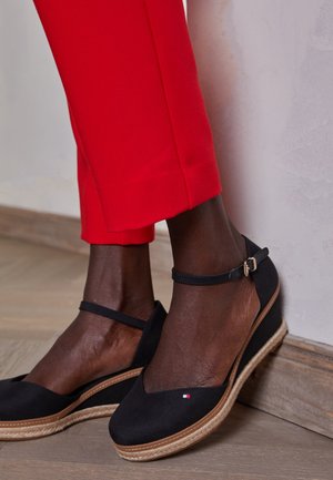 Esparteñas negras con cuña, con un frontal en forma de V, tira en el tobillo y suela de yute texturizada. Combinadas con pantalones rojos ajustados frente a una pared neutra.