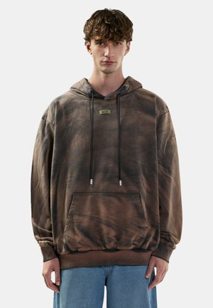 Giovane uomo con capelli ricci che indossa una felpa con cappuccio marrone tie-dye con una tasca frontale e coulisse nere, abbinata a jeans blu.