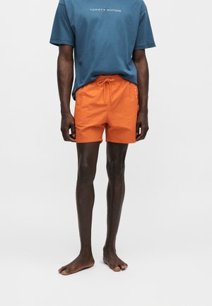 Άνδρας με μπλε μπλούζα Tommy Hilfiger και πορτοκαλί σορτς με κορδόνι, στέκεται ξυπόλητος σε απλό ανοιχτό φόντο.