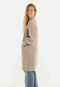 Vrouw met lang golvend haar, gekleed in een beige gebreide cardigan met zakken en lichte spijkerbroek, glimlachend en kijkend naar de zijkant.