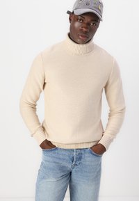 Maglione beige a collo alto con polsini e orlo a coste, abbinato a jeans azzurri. Il tessuto appare morbido e lavorato a maglia, offrendo un look informale.