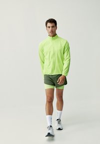 Giacca leggera con zip di un verde neon abbinata a pantaloni corti verde scuro con un accento giallo, calze sportive bianche e scarpe da corsa grigie.