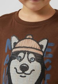 Brązowy bawełniany T-shirt z grafiką wilka w beżowej czapce, czarnymi konturami i niebieskim napisem. Wzór ma teksturowany, wypukły efekt.