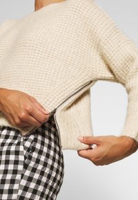 Pull crème texturé avec un détail de fermeture éclair latérale, doté d'une coupe décontractée et de poignets côtelés, associé à un bas à motif à carreaux noir et blanc.